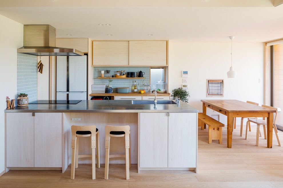 Inspiration för moderna linjära kök med öppen planlösning, med en integrerad diskho, släta luckor, skåp i ljust trä, bänkskiva i rostfritt stål, blått stänkskydd, vita vitvaror, ljust trägolv, en halv köksö och beiget golv