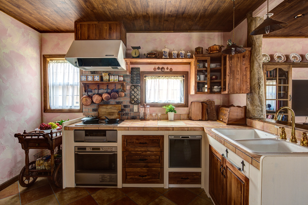 Modelo de cocinas en L rural con fregadero de doble seno, armarios con paneles empotrados, puertas de armario con efecto envejecido, encimera de azulejos, suelo marrón y encimeras naranjas