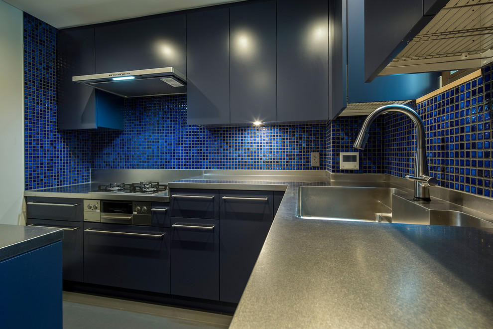Cette photo montre une cuisine américaine tendance en L avec un placard à porte plane, des portes de placard bleues, un évier intégré, un plan de travail en inox, une crédence en céramique, un sol en linoléum et un sol gris.
