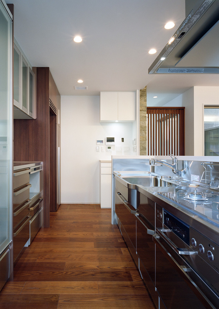 Asiatisk inredning av ett kök, med en integrerad diskho, släta luckor, bänkskiva i rostfritt stål, mellanmörkt trägolv, en halv köksö och brunt golv