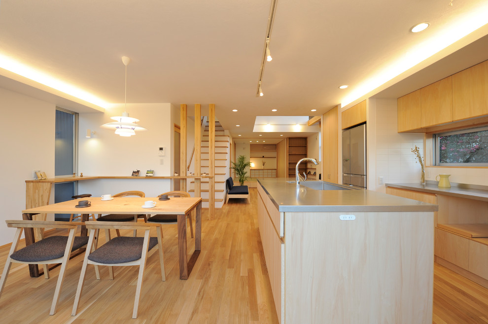 Photo of a scandi galley open plan kitchen in Other with an integrated sink, flat-panel cabinets, light wood cabinets, stainless steel worktops, white splashback, light hardwood flooring, an island, brown floors and beige worktops.