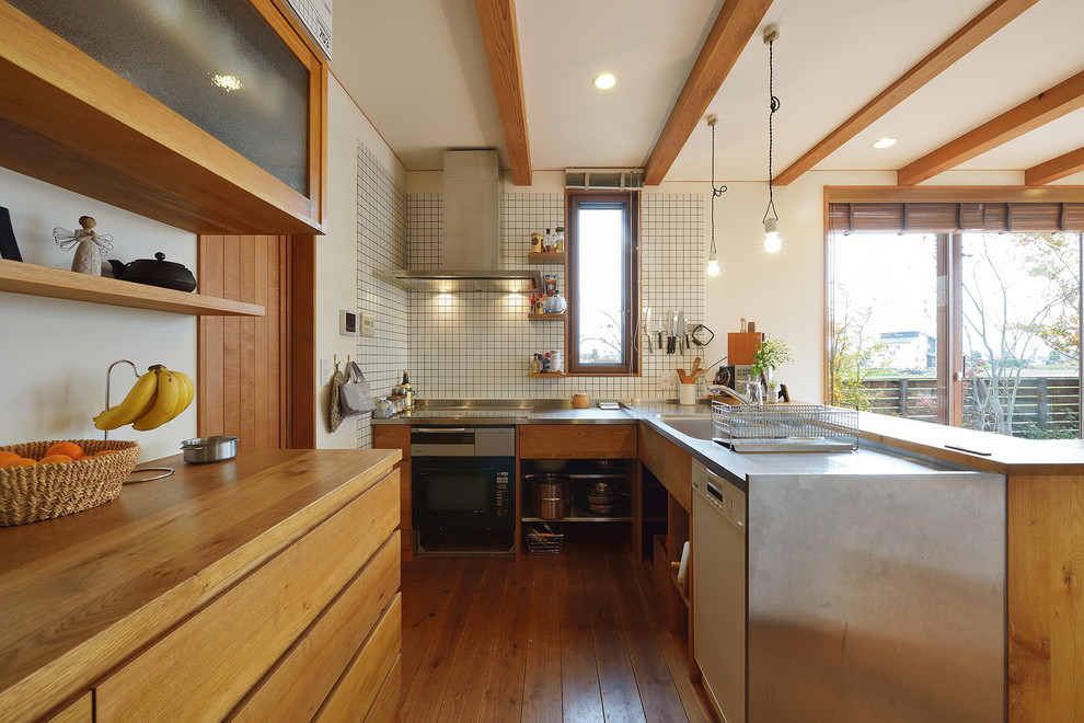 Foto på ett industriellt brun l-kök, med en enkel diskho, släta luckor, skåp i mellenmörkt trä, bänkskiva i rostfritt stål, vitt stänkskydd, stänkskydd i keramik, mellanmörkt trägolv, en halv köksö och brunt golv