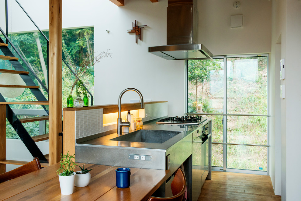Modern inredning av ett grå linjärt grått kök, med släta luckor, skåp i rostfritt stål, bänkskiva i rostfritt stål, vitt stänkskydd, stänkskydd i porslinskakel, brunt golv, en integrerad diskho och mellanmörkt trägolv
