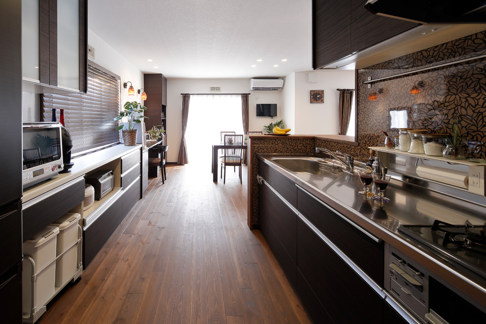 Example of a galley medium tone wood floor and brown floor open concept kitchen design in Other with a single-bowl sink, flat-panel cabinets, black cabinets, stainless steel countertops, brown backsplash and a peninsula