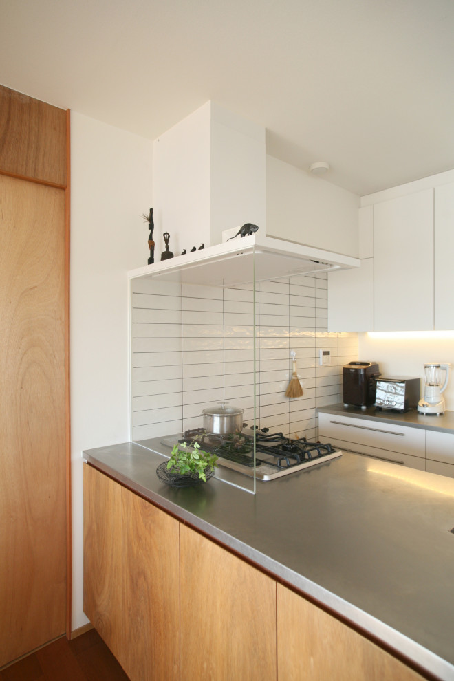 Foto på ett litet funkis grå kök, med en undermonterad diskho, släta luckor, vita skåp, bänkskiva i rostfritt stål, vitt stänkskydd, stänkskydd i porslinskakel, svarta vitvaror, mörkt trägolv, en halv köksö och brunt golv