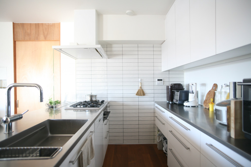 Inredning av ett modernt litet grå grått kök, med en undermonterad diskho, släta luckor, vita skåp, bänkskiva i rostfritt stål, vitt stänkskydd, stänkskydd i porslinskakel, svarta vitvaror, mörkt trägolv, en halv köksö och brunt golv