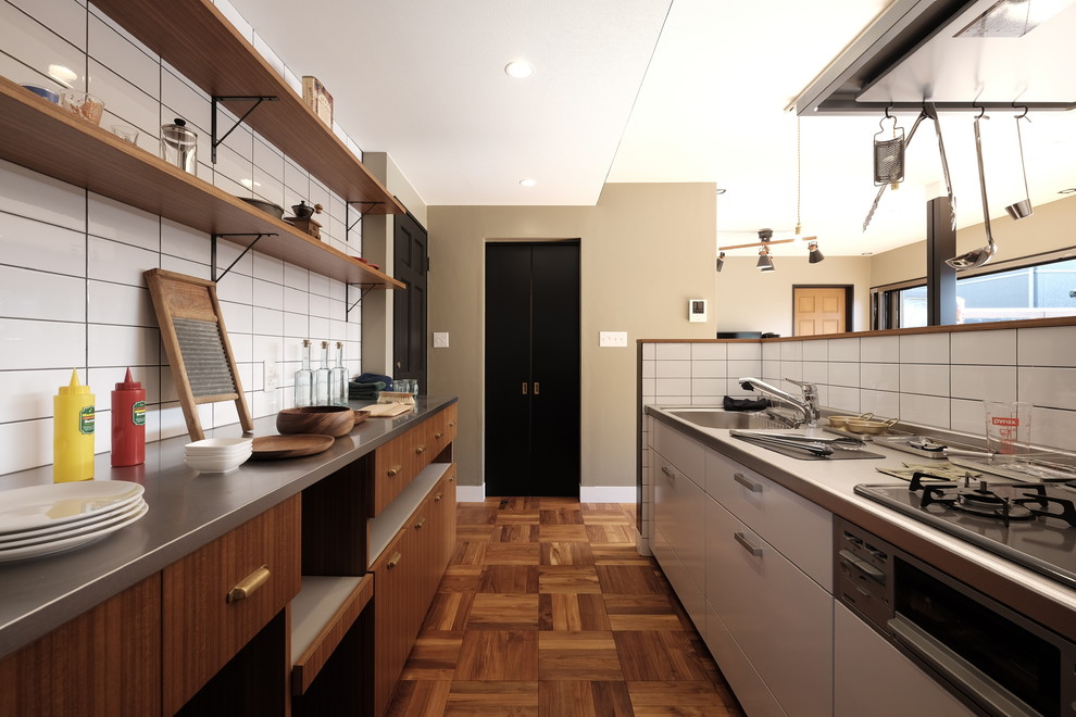 他の地域にあるインダストリアルスタイルのおしゃれなキッチン (シングルシンク、フラットパネル扉のキャビネット、中間色木目調キャビネット、ステンレスカウンター、白いキッチンパネル、サブウェイタイルのキッチンパネル、無垢フローリング、茶色い床) の写真
