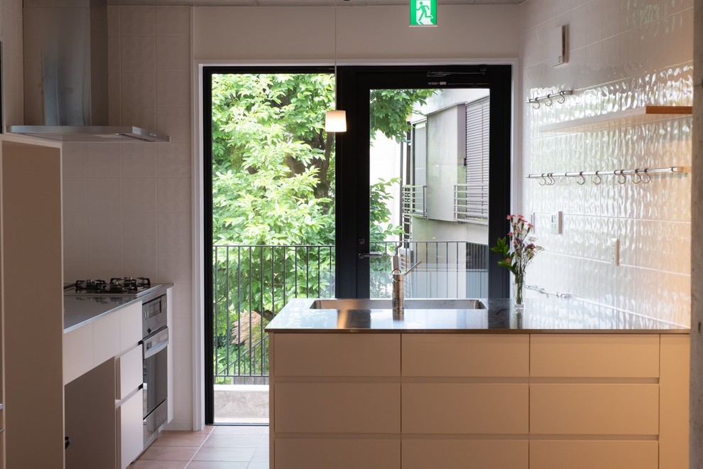 Trendy terra-cotta tile and brown floor kitchen photo in Tokyo with an integrated sink, flat-panel cabinets, white cabinets, stainless steel countertops, white backsplash, ceramic backsplash, white appliances, a peninsula and gray countertops