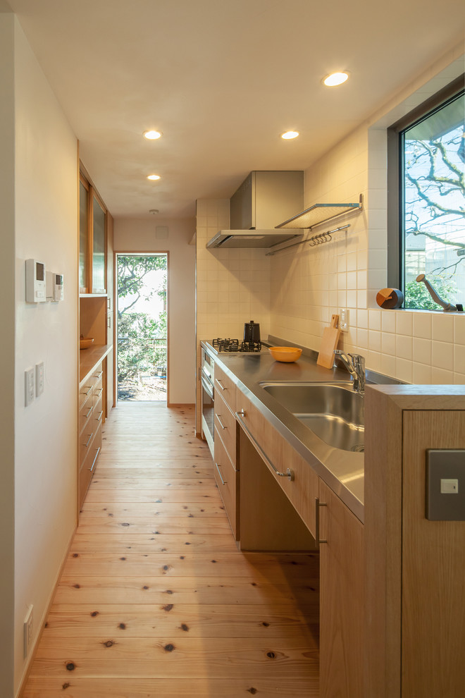 Bild på ett litet funkis linjärt kök med öppen planlösning, med en integrerad diskho, släta luckor, skåp i ljust trä, bänkskiva i rostfritt stål, vitt stänkskydd, rostfria vitvaror, ljust trägolv, stänkskydd i porslinskakel, en köksö och brunt golv