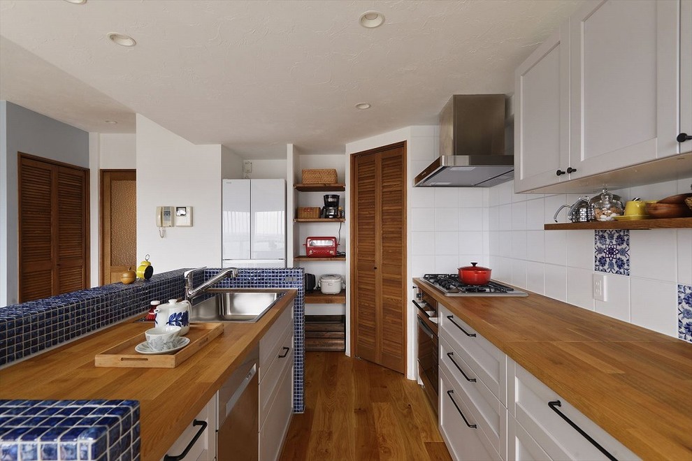 地中海スタイルのおしゃれなキッチン (シングルシンク、落し込みパネル扉のキャビネット、白いキャビネット、木材カウンター、青いキッチンパネル、セラミックタイルのキッチンパネル、無垢フローリング、茶色い床、茶色いキッチンカウンター) の写真