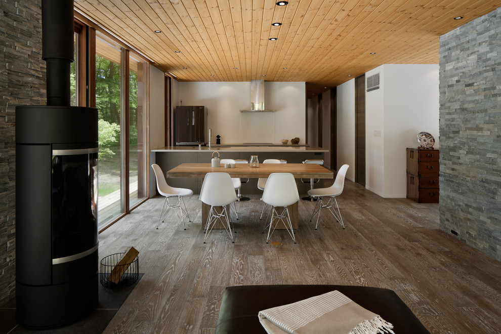 Immagine di una sala da pranzo aperta verso il soggiorno minimalista con pareti bianche, pavimento in legno verniciato, pavimento marrone, stufa a legna e cornice del camino in pietra