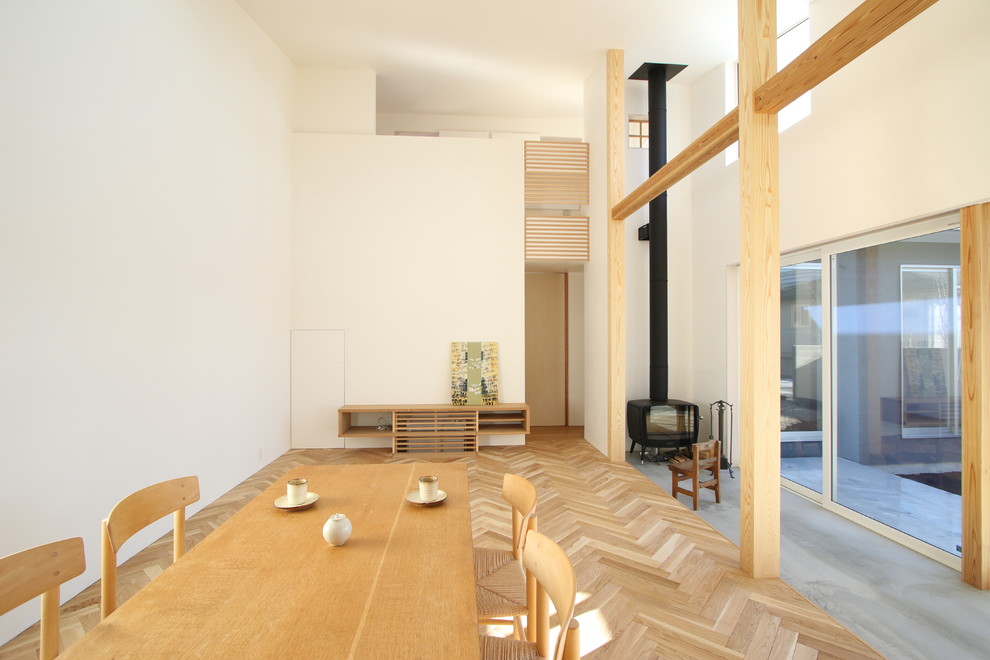 Imagen de comedor minimalista con paredes blancas, suelo de madera clara, estufa de leña, marco de chimenea de hormigón y suelo beige