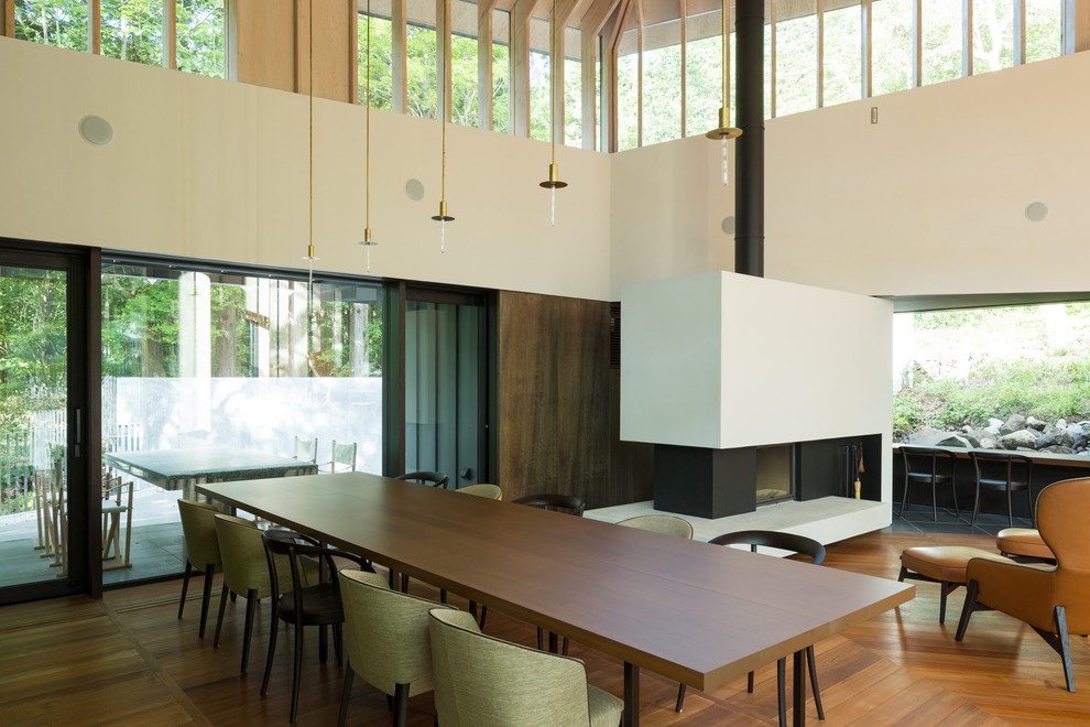 Imagen de comedor minimalista abierto con paredes blancas, suelo de madera en tonos medios, chimenea de doble cara y marco de chimenea de yeso