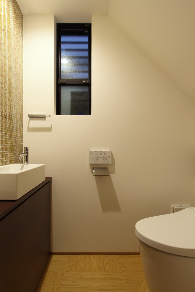 Foto på ett litet orientaliskt brun toalett, med släta luckor, bruna skåp, en toalettstol med hel cisternkåpa, brun kakel, glasskiva, vita väggar, ett fristående handfat och beiget golv