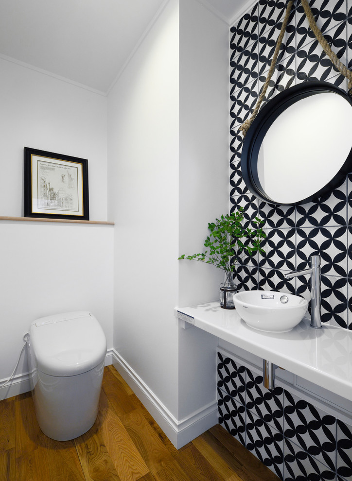 Bild på ett vintage vit vitt toalett, med en toalettstol med hel cisternkåpa, svart och vit kakel, vita väggar, mellanmörkt trägolv, ett fristående handfat och brunt golv