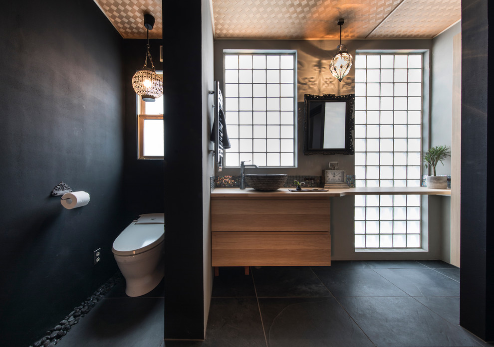 Пример оригинального дизайна: туалет в современном стиле с плоскими фасадами, фасадами цвета дерева среднего тона, черными стенами, настольной раковиной, столешницей из дерева, черным полом и коричневой столешницей