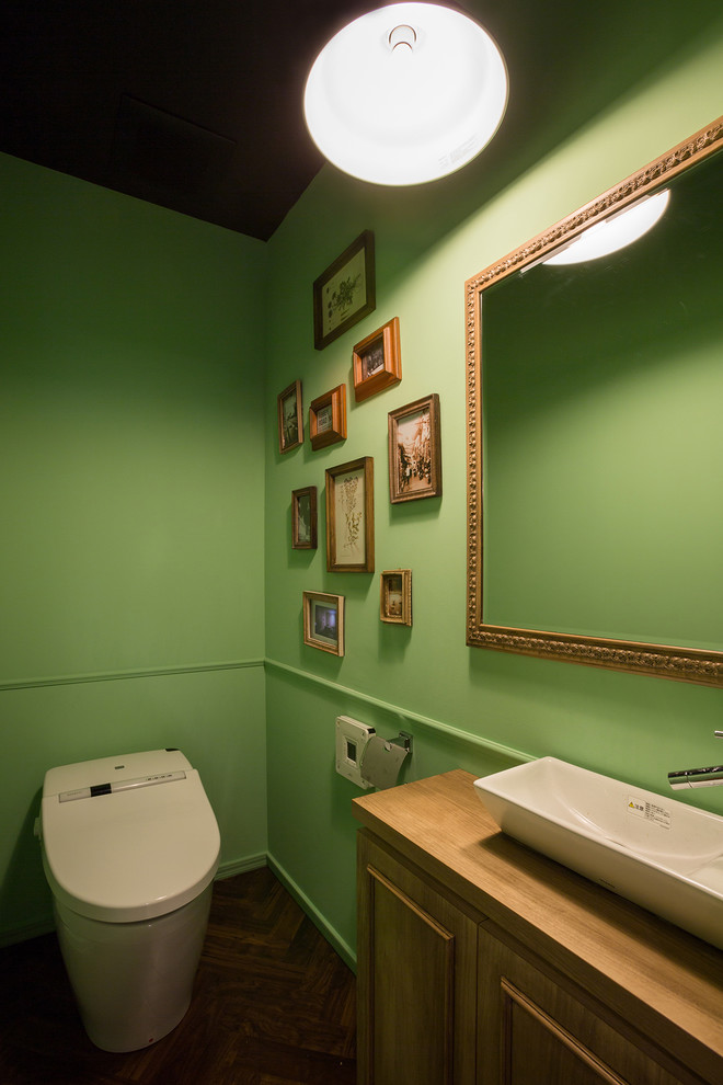 Mittelgroße Klassische Gästetoilette mit grüner Wandfarbe, Aufsatzwaschbecken, Waschtisch aus Holz, Schrankfronten mit vertiefter Füllung, hellen Holzschränken, brauner Waschtischplatte, Toilette mit Aufsatzspülkasten, Laminat und braunem Boden in Yokohama