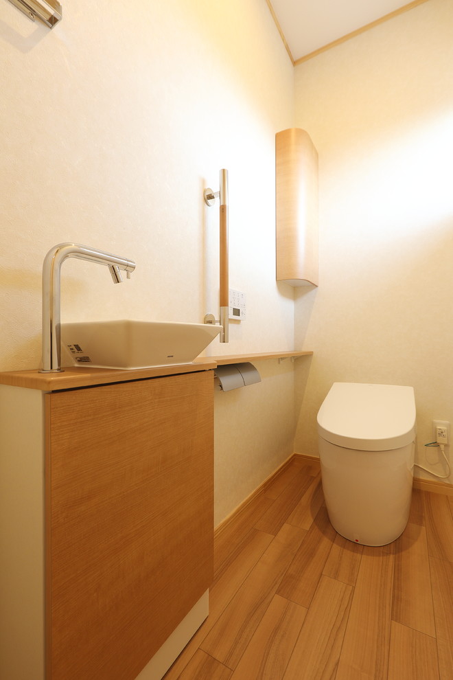 Пример оригинального дизайна: туалет среднего размера с светлыми деревянными фасадами, унитазом-моноблоком, белыми стенами, полом из фанеры, настольной раковиной, бежевым полом и бежевой столешницей