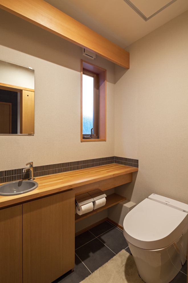 東京都下にある和風のおしゃれなトイレ・洗面所 (フラットパネル扉のキャビネット、中間色木目調キャビネット、ベージュの壁、オーバーカウンターシンク、木製洗面台、黒い床) の写真