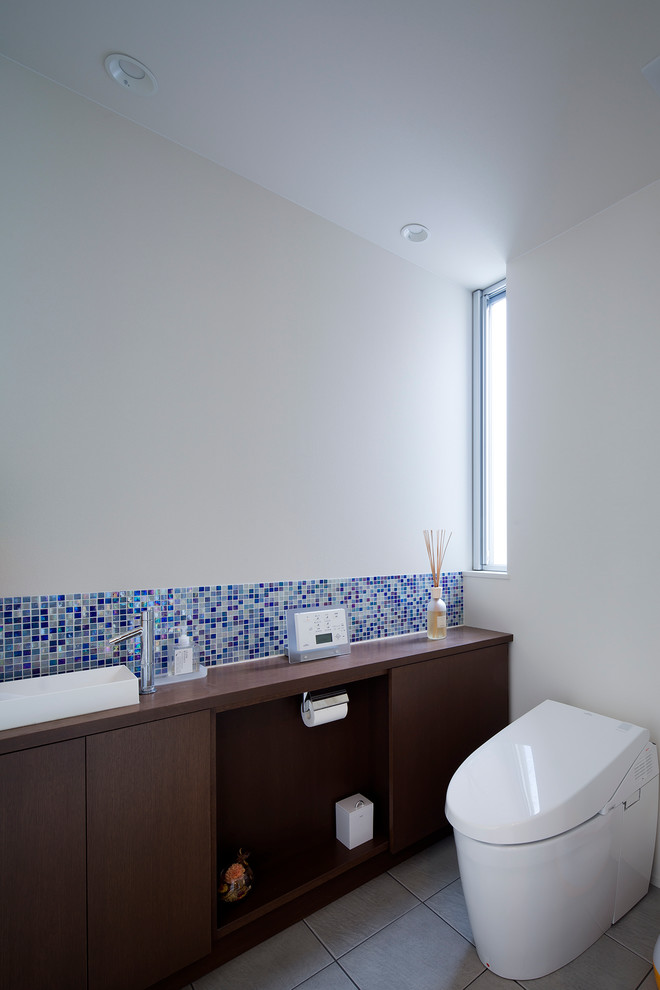 Modern inredning av ett brun brunt toalett, med en toalettstol med hel cisternkåpa, vita väggar, släta luckor, skåp i mörkt trä, blå kakel, ett fristående handfat och träbänkskiva