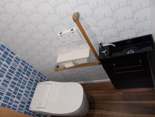 Bild på ett funkis toalett, med en toalettstol med hel cisternkåpa, blå kakel, vita väggar, plywoodgolv och brunt golv