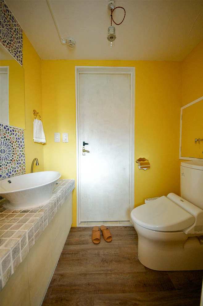 Medelhavsstil inredning av ett toalett, med grå kakel, flerfärgad kakel, gula väggar, mellanmörkt trägolv, ett fristående handfat, kaklad bänkskiva och en toalettstol med separat cisternkåpa