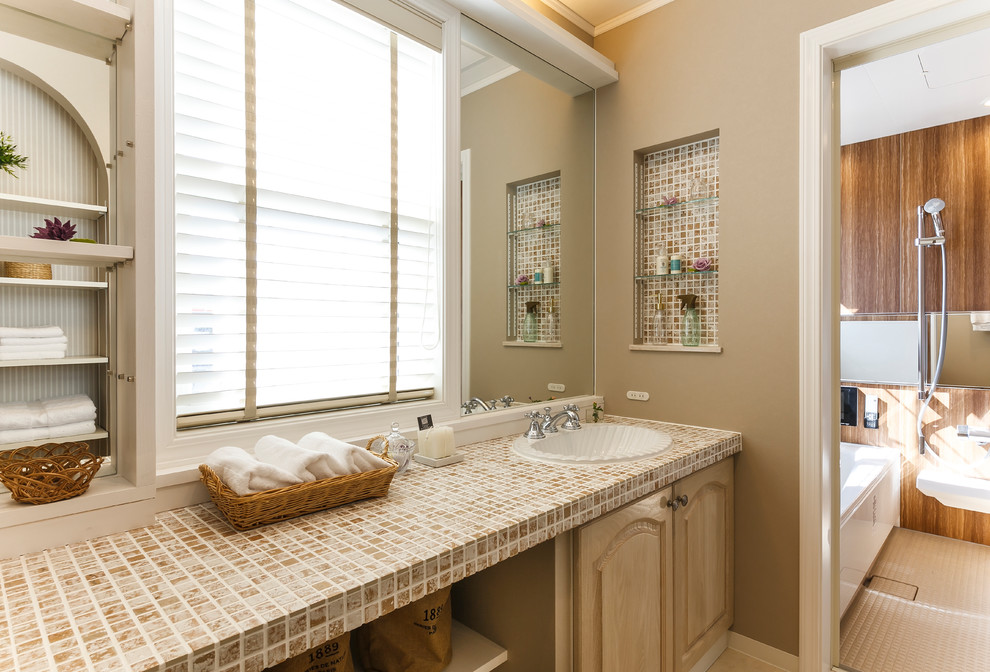 Example of a trendy beige floor powder room design in Other with recessed-panel cabinets, light wood cabinets, brown walls, a drop-in sink, tile countertops and beige countertops