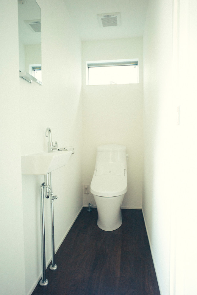 Cette image montre un WC et toilettes minimaliste de taille moyenne avec un placard à porte plane, des portes de placard blanches, WC séparés, un mur blanc, parquet foncé et un lavabo suspendu.