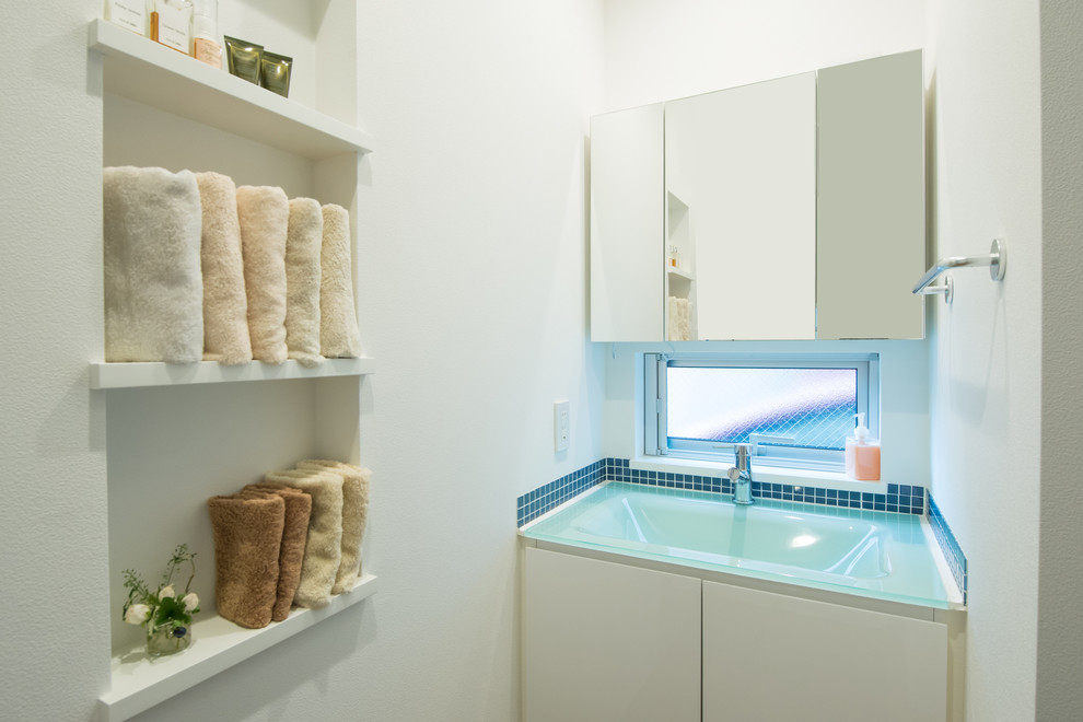 Kleine Gästetoilette mit flächenbündigen Schrankfronten, weißen Schränken, blauen Fliesen, weißer Wandfarbe, integriertem Waschbecken und Glaswaschbecken/Glaswaschtisch in Tokio