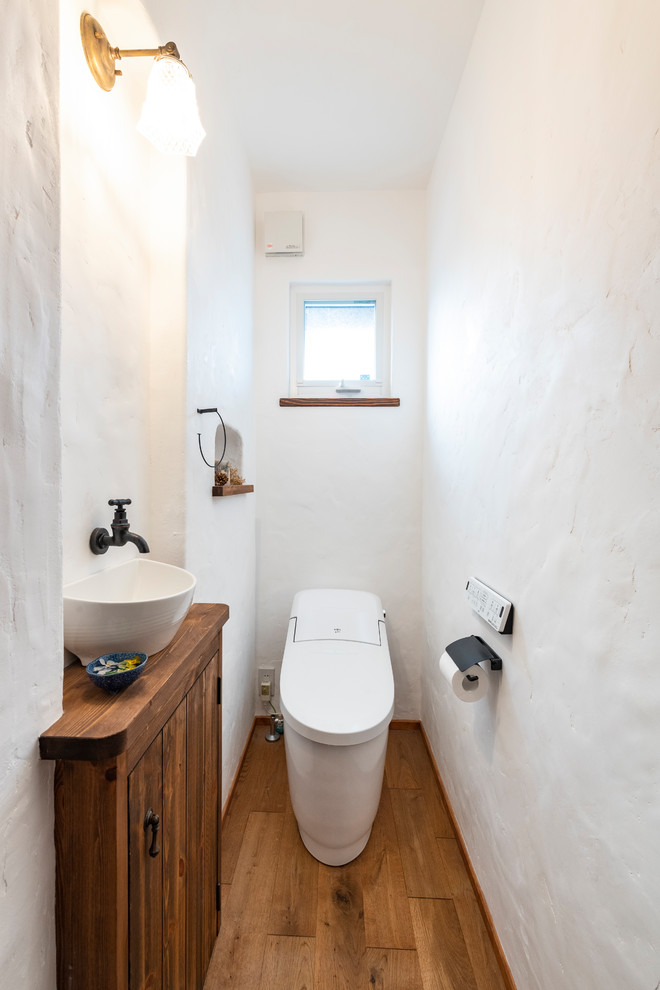 他の地域にある小さな地中海スタイルのおしゃれなトイレ・洗面所 (ヴィンテージ仕上げキャビネット、一体型トイレ	、白い壁、濃色無垢フローリング、オーバーカウンターシンク、茶色い床) の写真