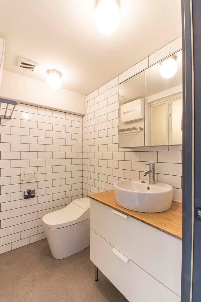 Bild på ett minimalistiskt brun brunt toalett, med släta luckor, vita skåp, vit kakel, tunnelbanekakel, vita väggar, ett fristående handfat, träbänkskiva och grått golv