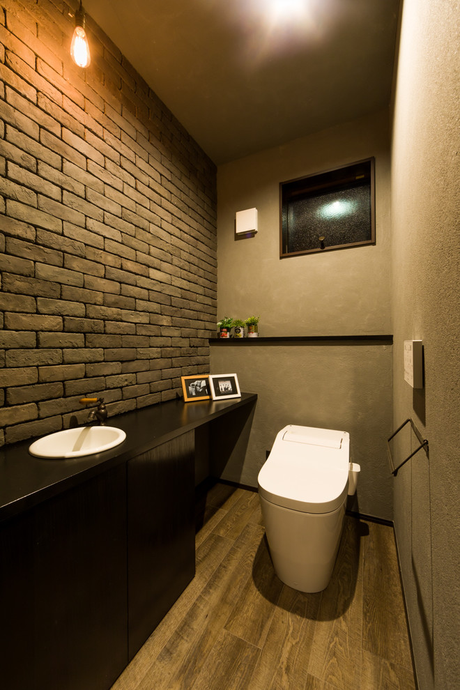 Foto på ett funkis svart toalett, med svarta skåp, en toalettstol med hel cisternkåpa, brun kakel, cementkakel, grå väggar, mörkt trägolv och brunt golv