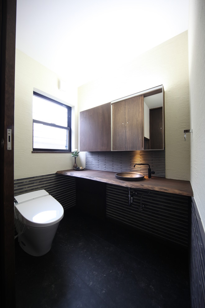 Cette photo montre un WC et toilettes asiatique de taille moyenne avec un placard en trompe-l'oeil, des portes de placard marrons, WC à poser, un carrelage gris, un mur beige, un sol en carrelage de porcelaine, un lavabo posé, un plan de toilette en bois, un sol noir et un plan de toilette marron.