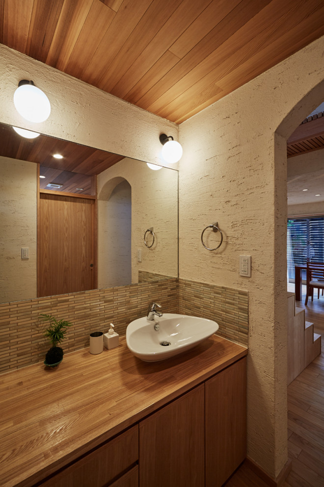 Пример оригинального дизайна: туалет в восточном стиле с плоскими фасадами, светлыми деревянными фасадами, унитазом-моноблоком, зеленой плиткой, керамогранитной плиткой, бежевыми стенами, светлым паркетным полом, консольной раковиной, столешницей из дерева, бежевым полом, бежевой столешницей, встроенной тумбой и деревянным потолком