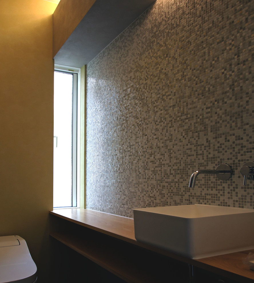 Inspiration pour un WC et toilettes minimaliste avec un placard sans porte, des portes de placard marrons, WC à poser, un carrelage gris, mosaïque, un mur beige, un sol en vinyl, un lavabo posé, un plan de toilette en bois, un sol gris et un plan de toilette marron.