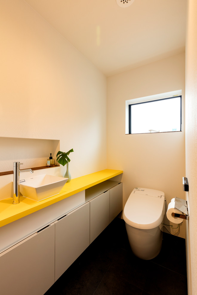 На фото: туалет среднего размера в стиле модернизм с белыми фасадами, унитазом-моноблоком, белыми стенами, накладной раковиной, столешницей из искусственного камня, черным полом, желтой столешницей, потолком с обоями и обоями на стенах