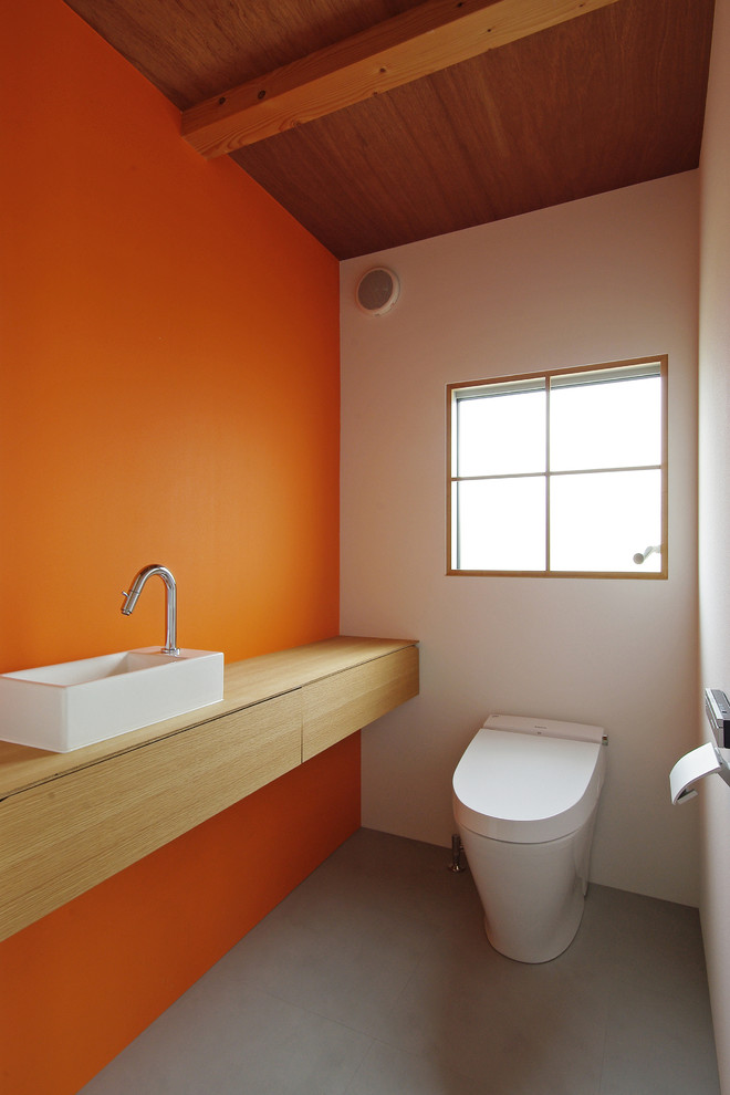 Foto på ett funkis beige toalett, med släta luckor, skåp i ljust trä, en vägghängd toalettstol, orange väggar, betonggolv, ett fristående handfat, träbänkskiva och grått golv