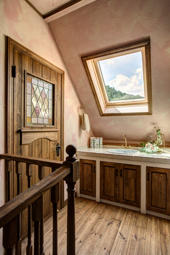 Photo of a traditional cloakroom in Other with recessed-panel cabinets, distressed cabinets, pink walls, medium hardwood flooring, a built-in sink, tiled worktops, brown floors and beige worktops.