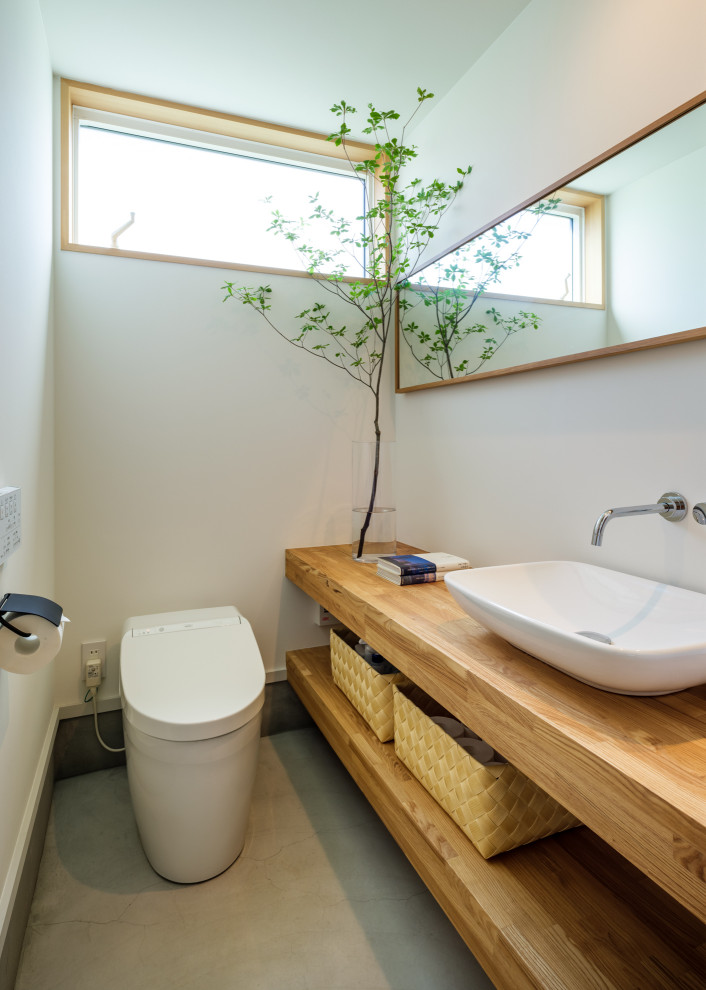 Bild på ett orientaliskt brun brunt toalett, med öppna hyllor, skåp i mellenmörkt trä, en toalettstol med hel cisternkåpa, vita väggar, betonggolv, ett fristående handfat, träbänkskiva och grått golv