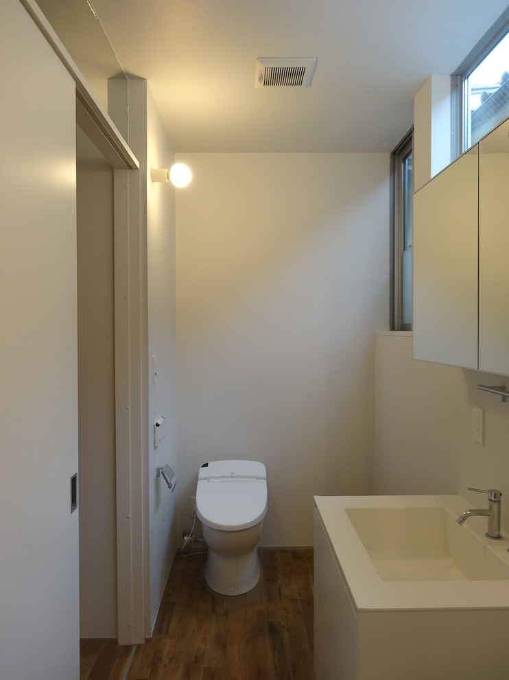 東京23区にある低価格の中くらいなモダンスタイルのおしゃれなトイレ・洗面所 (フラットパネル扉のキャビネット、白いキャビネット、一体型トイレ	、白いタイル、白い壁、濃色無垢フローリング、一体型シンク、人工大理石カウンター、茶色い床、白い洗面カウンター) の写真