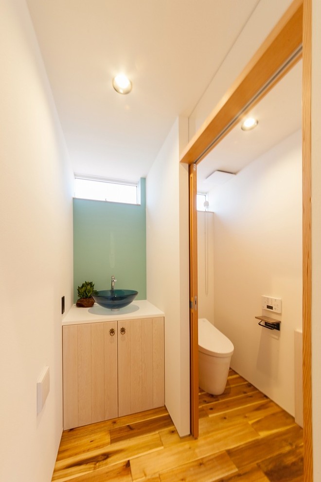 Idées déco pour un WC et toilettes asiatique avec un placard à porte plane, des portes de placard beiges, un mur blanc, un sol en bois brun, une vasque et un sol marron.
