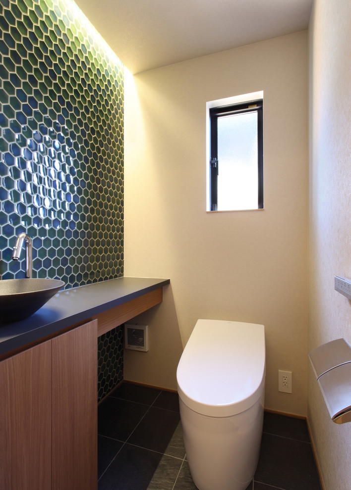 Bild på ett litet orientaliskt svart svart toalett, med släta luckor, bruna skåp, en toalettstol med hel cisternkåpa, grön kakel, mosaik, vita väggar, klinkergolv i keramik, ett fristående handfat, laminatbänkskiva och svart golv