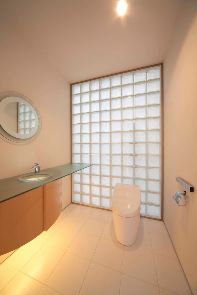 Exemple d'un WC et toilettes asiatique en bois clair avec un placard à porte plane, un mur blanc, un plan de toilette en verre et un sol blanc.