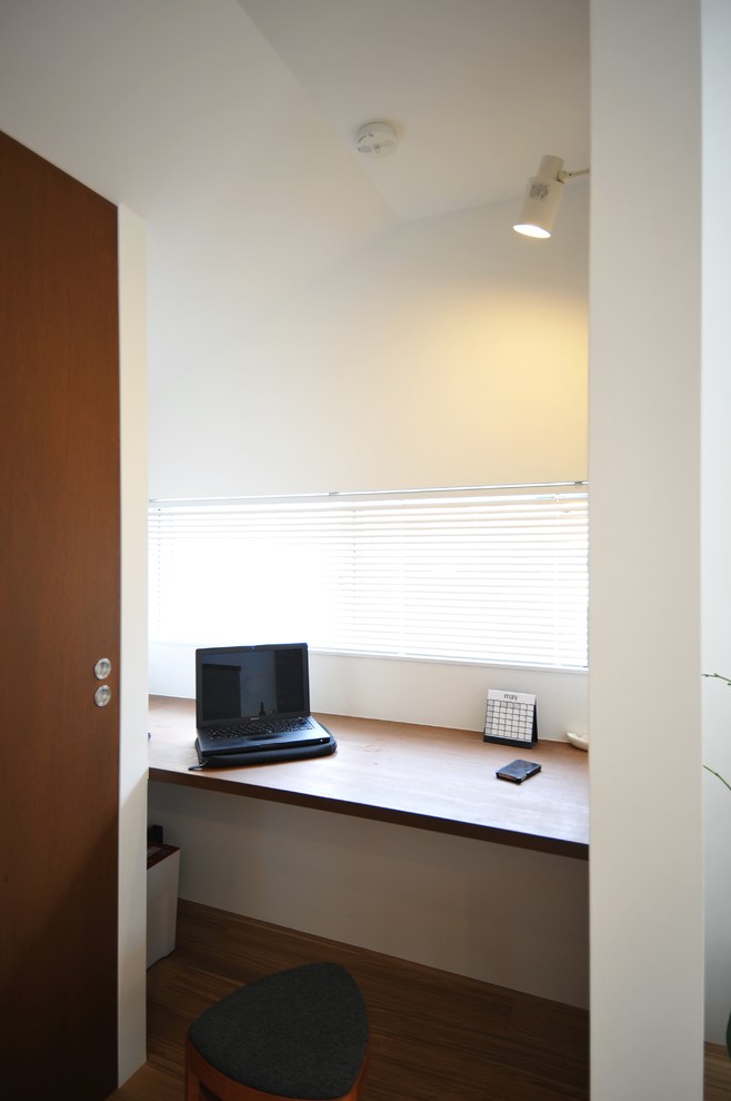 Ejemplo de despacho actual pequeño con biblioteca, paredes blancas, suelo de contrachapado, escritorio empotrado y suelo marrón