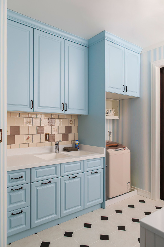Modern inredning av en stor linjär tvättstuga enbart för tvätt, med en dubbel diskho, luckor med upphöjd panel, blå skåp, beige väggar, klinkergolv i keramik och tvättmaskin och torktumlare byggt in i ett skåp