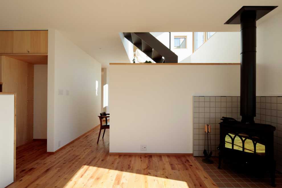На фото: открытая гостиная комната среднего размера в скандинавском стиле с белыми стенами, полом из фанеры, печью-буржуйкой, фасадом камина из плитки, коричневым полом и телевизором на стене с