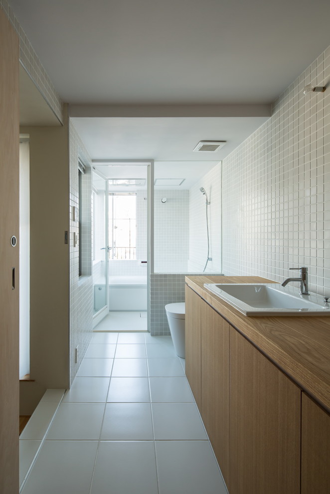 Modelo de cuarto de baño actual con armarios con paneles lisos, puertas de armario de madera clara, sanitario de una pieza, baldosas y/o azulejos blancos, paredes blancas, lavabo encastrado y encimera de madera