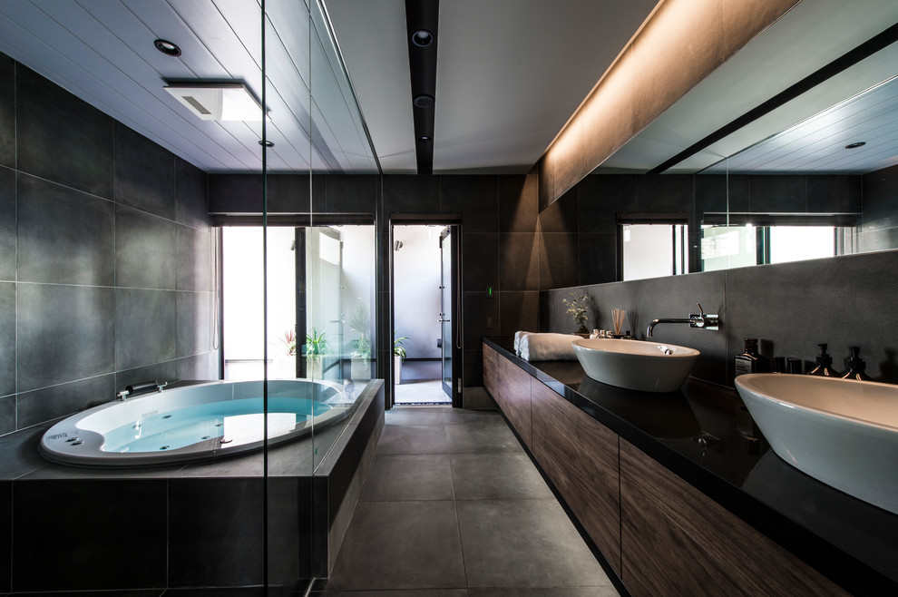 Bild på ett funkis badrum, med släta luckor, skåp i mörkt trä, ett platsbyggt badkar, grå kakel, grå väggar, ett fristående handfat och grått golv