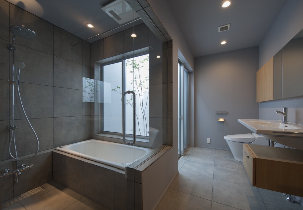 Photo of a modern ensuite bathroom in Tokyo with flat-panel cabinets, light wood cabinets, a built-in bath, a double shower, a one-piece toilet, grey tiles, grey walls, a wall-mounted sink, grey floors, an open shower, ceramic flooring and white worktops.