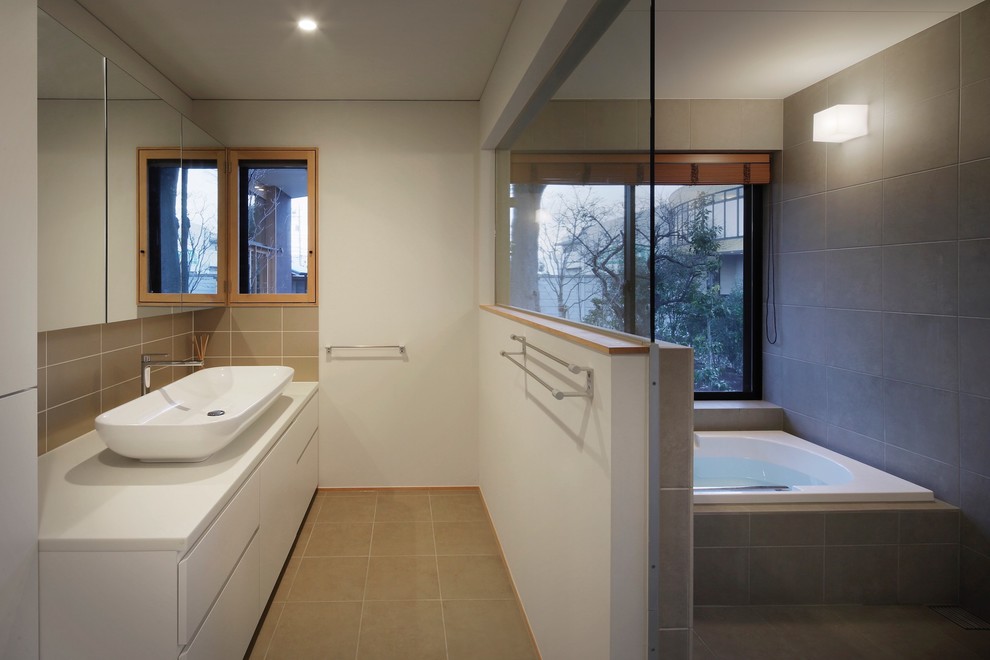 На фото: большая ванная комната в стиле модернизм с плоскими фасадами, белыми фасадами, накладной ванной, душевой комнатой, унитазом-моноблоком, серой плиткой, керамической плиткой, белыми стенами, полом из керамической плитки, настольной раковиной, столешницей из искусственного камня, бежевым полом, открытым душем и белой столешницей